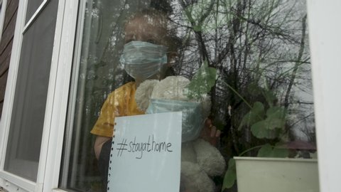 Stay at home quarantine for coronavirus pandemic prevention. Child and his teddy bear both in protective medical masks sits on windowsill and looks out window. View from street. Prevention epidemic