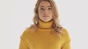 Pretty blonde woman in sweater becoming surprised and holding her cheeks over grey background