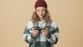 Cheerful pretty woman in knit hat and shirt playing on smartphone over beige background