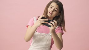 Cheerful pretty brunette woman in overalls playing on smartphone over pink background