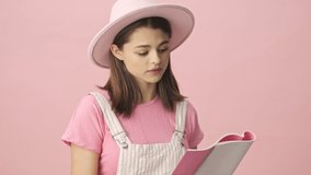 Calm pretty brunette woman in overalls and hat reading something in notebook and becoming happy over pink background
