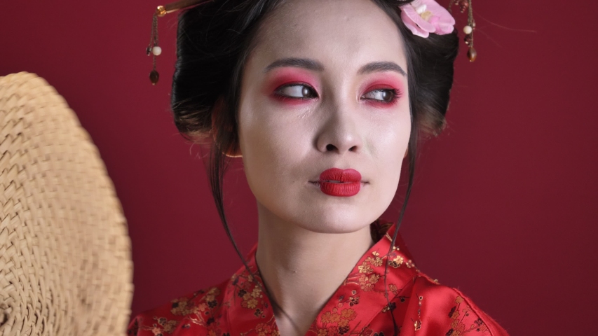 A close-up view of an displeased young geisha woman in traditional japanese kimono is fanning herself isolated over red background