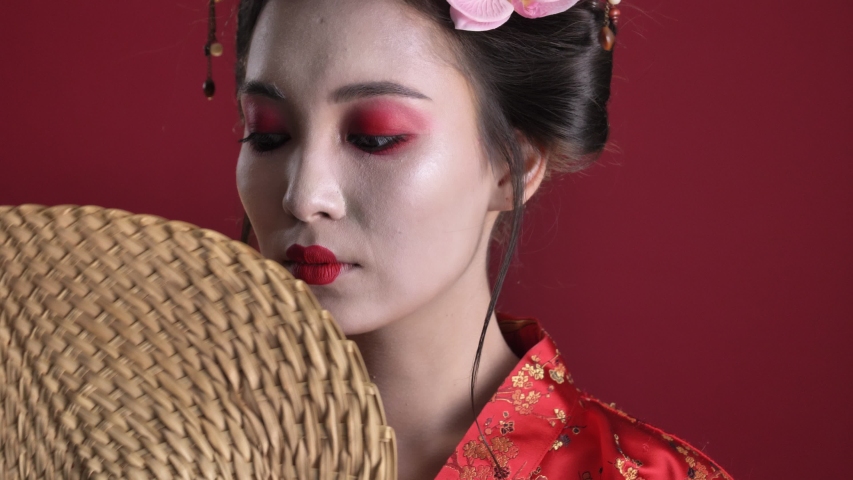 A close-up view of a beautiful young geisha woman in traditional japanese kimono is posing with wooden hand fan while moving it from one side to another isolated over red background
