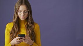 A calm nice teenage girl is using her smartphone standing isolated over a purple background in studio