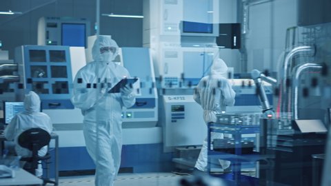 Research Factory Cleanroom: Engineer Wearing Coverall and Mask, Walking Through Workshopop Uses Tablet Computer. In Background Professionals Work on Modern High Tech CNC Machinery