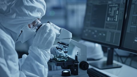 Research Factory Cleanroom: Engineer / Scientist wearing Coverall and Gloves Uses Microscope to Inspect Samples, Developing High Tech Modern Technology for Medical and Precision Electronics Industry