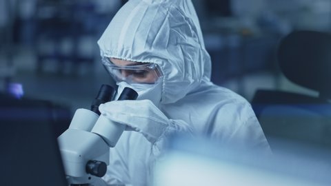 Research Factory Cleanroom: Engineer / Scientist wearing Coverall and Gloves Uses Microscope to Inspect Samples, Developing High Tech Modern Electronics for Medical,High Precision Electronics Industry