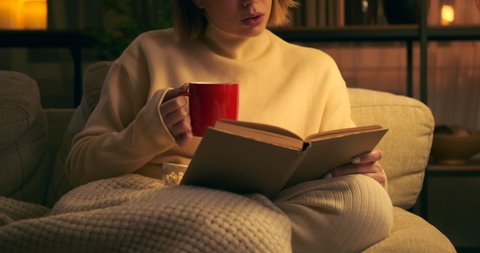 Woman drinking coffee and reading book on couch at night