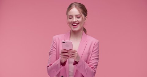 Extremely Happy, Satisfied Girl is using her Smartphone isolated on pink Studio. Funny Smingly Girl is texting Messages, playing with her Mobile Phone. Having nice pink Outfit and Phone Case. Apps.