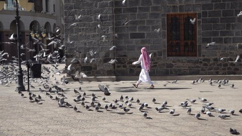 MEDINA, SAUDI ARABIA – DECEMBER 2019: Boy in traditional Saudi dress walks through flock of pigeons in central Medina, religion and traditional culture in the Middle East

