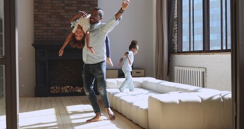 Carefree young african american father dancing barefoot on heated wooden floor with cute daughter on shoulder. Happy biracial man having fun with small adorable children, playing together at home.