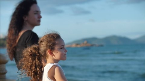 Happy European family of two mother and daughter stand on the beach at sunset, look into the distance at sea or ocean. Slow motion. Family, vacation and travel concept.