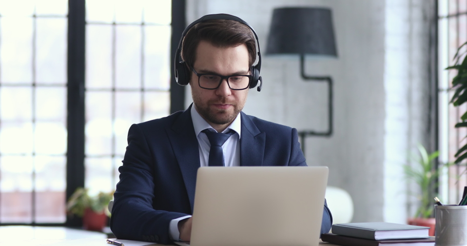 Confident businessman wears suit and headset makes conference video call on laptop. Male call center agent, customer support manager or telemarketer communicating in webcam chat on computer in office. Royalty-Free Stock Footage #1048286560