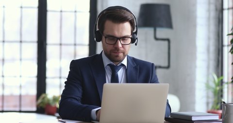 Confident businessman wears suit and headset makes conference video call on laptop. Male call center agent, customer support manager or telemarketer communicating in webcam chat on computer in office.
