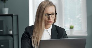 Close up of beautiful mature woman in eyeglasses and black suit working on laptop with serious facial expression. Female manager with blond hair professionaly doing her job