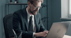 Responsible businessman in formal clothing and eyeglasses sitting at table at private office and working on laptop. Bearded man using computer at working place.