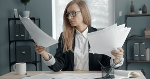 Concentrated middle-aged woman in black suit and eyewear sitting at office and searching for important documents among other papers lying on table. Concept of working process and business