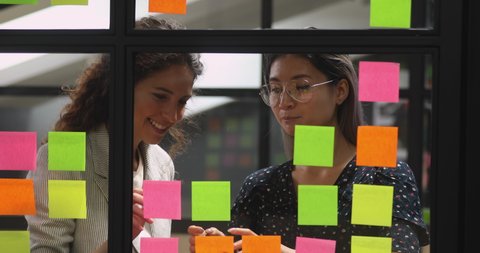 Positive multiethnic young businesswomen managing working process together at glass kanban board, using colorful paper stickers. Smart female diverse colleagues discussing project tasks in office.