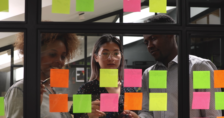 Happy mixed race young colleagues working together near window kanban board in modern office. Smiling diverse project managers managing workflow, discussing ideas, writing notes on paper stickers. Royalty-Free Stock Footage #1048307419