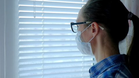 A woman at home looks out the window in a medical mask, quarantined due to the spread of coronavirus