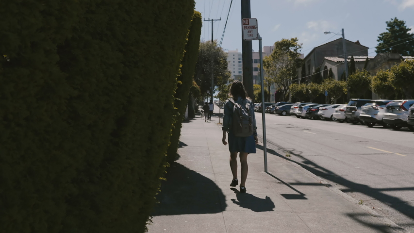 Camera follows happy young tourist woman with backpack walking along summer San Francisco street touching green fence.