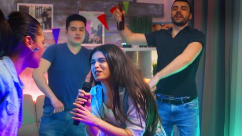 Beautiful young woman singing a song on microphone while partying with her cheerful friends. Karaoke at wild college party in room with neon lights, disco ball and alcohol.