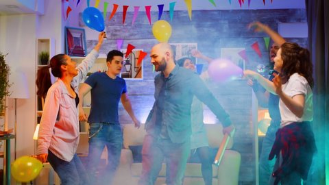 Attractive young man throwing confetti while partying with his group of friends in a room with neon lights, disco ball and alcohol. Slow motion shot