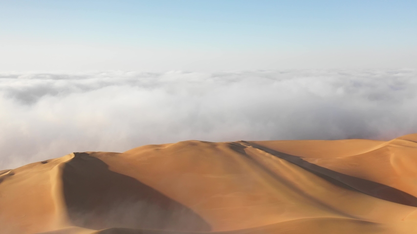 Aerial view of a drone flying over massive sand dunes covered by thick fog clouds at sunrise. Liwa desert, Abu Dhabi, United Arab Emirates. Royalty-Free Stock Footage #1048349581