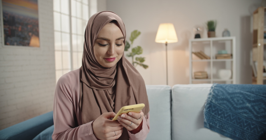 Young muslim girl wearing traditional hijab scarf looking at her smartphone. Successful female freelance manager or university student working at home or office 4k footage Royalty-Free Stock Footage #1048352209