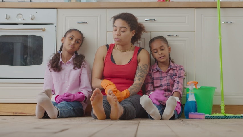 Tired pretty african american mother and adorable preadolescent daughters sitting on kitchen floor with cleaning supplies after finishing cleanup in domestic kitchen, feeling exhausted and happy.