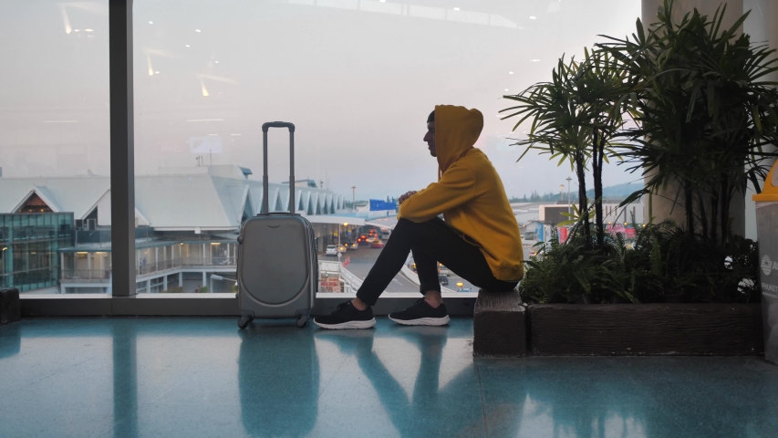Young hooded man tourist with luggage waiting for boarding at the airport terminal sitting near window