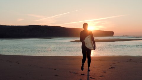 Revealing shot of a young woman surfer with a blonde hair wearing a wetsuit and holding surfboard running towards the ocean with waves on the golden sunset / sunrise. Sun is making a red colour. 