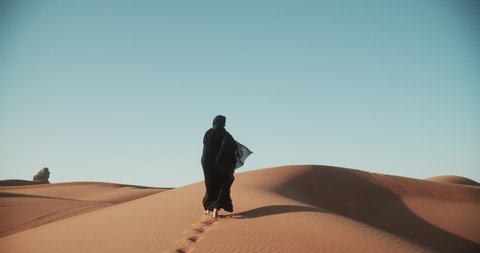 Young beautiful girl in traditional Abaya dress walks barefoot through the dunes in the desert of Dubai. 4K Slow Motion