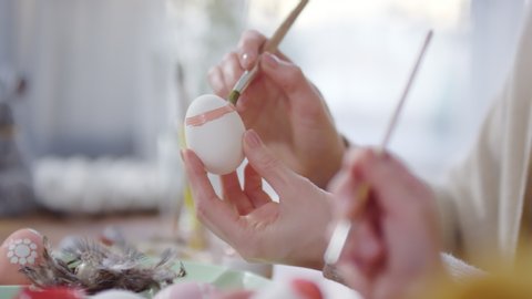 Handheld close up shot of hands of unrecognizable woman and girl using brushes and painting on white boiled eggs while preparing for Easter