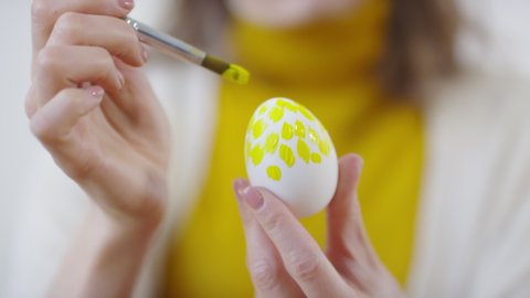 Close up shot of unrecognizable woman using brush and painting yellow pattern on white Easter egg