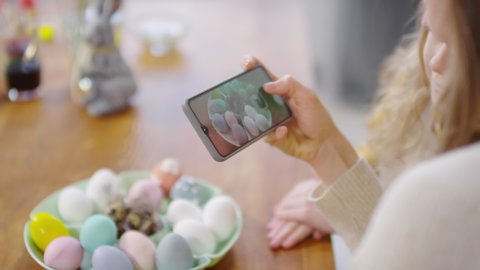 Handheld close up of unrecognizable woman holding mobile phone and taking photos of tray of Easter eggs as her cute daughter sitting beside her and watching
