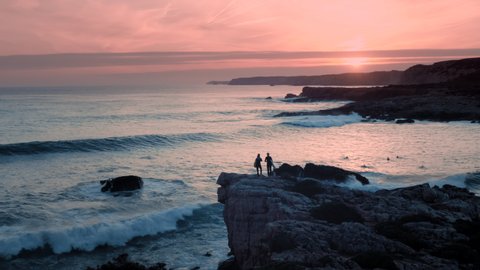 Beautiful aerial shot of two female surfers standing at the edge of cliff / rock and watching waves at sunset. Use for fitness/lifestyle advertising / commercial.