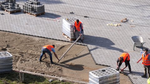 Construction workers are installing interlocking pavers. Two man are leveling the sand bedding with a screed board while others are laying pavers in place in Cluj-Napoca, Romania on March 14, 2020