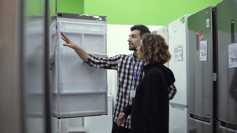 Young woman standing in front the open door of refrigerator with male consultant discussing design and quality before buying in a consumer electronics store. Discussing characteristics with store