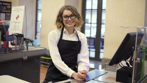 Woman in black apron as a cashier at the cash register in the supermarket or discounter. Smiling short haired curly woman smiling to the camera