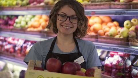 Beautiful smiling young female supermarket employee in black apron holding a box full of apples in front of shelf in supermarket with pretty face looking at camera professional front portrait startup