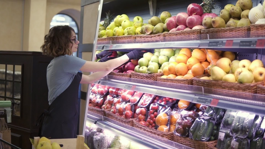 Close up of pretty caucasian worker in black apron and gloves stocking the fruits in supermarket. Young employee in glasses at work. Curly female arranging big red apples on shelf. Slow motion. Side Royalty-Free Stock Footage #1048376266