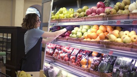 Close up of pretty caucasian worker in black apron and gloves stocking the fruits in supermarket. Young employee in glasses at work. Curly female arranging big red apples on shelf. Slow motion. Side