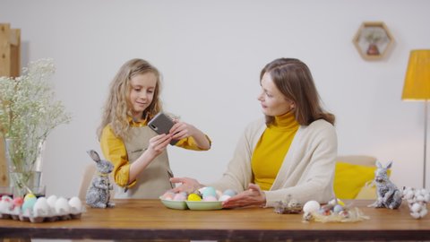 Handheld medium shot of beautiful young woman smiling and talking to cute girl taking photos of Easter eggs lying on tray. Bunny figurines and flowers in vase standing on table