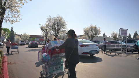 March 13, 2020: Woman wearing mask pushes shopping cart with toilet paper and other goods in Costco parking lot. Long line of people behind here trying to enter the store. 