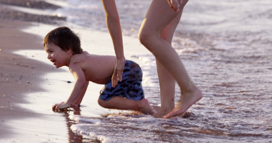 Toddler boy crawling on ocean beach stands up with mother - side profile
