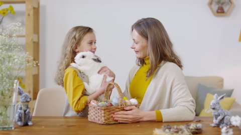 Handheld shot of happy young woman smiling and petting cute bunny held by her little daughter. Basket of Easter eggs standing on table