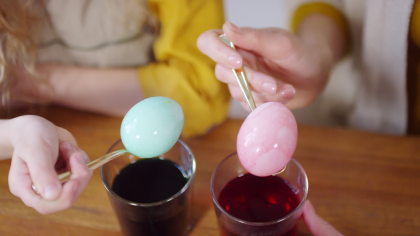 High angle close up of hands of unrecognizable girl and woman holding spoons with blue and red Easter eggs over glass cups filled with dye
