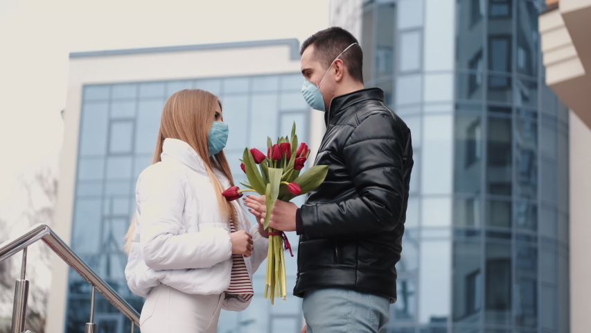 European couple in masks are standing together. The man is making a gift of flowers for a girl.