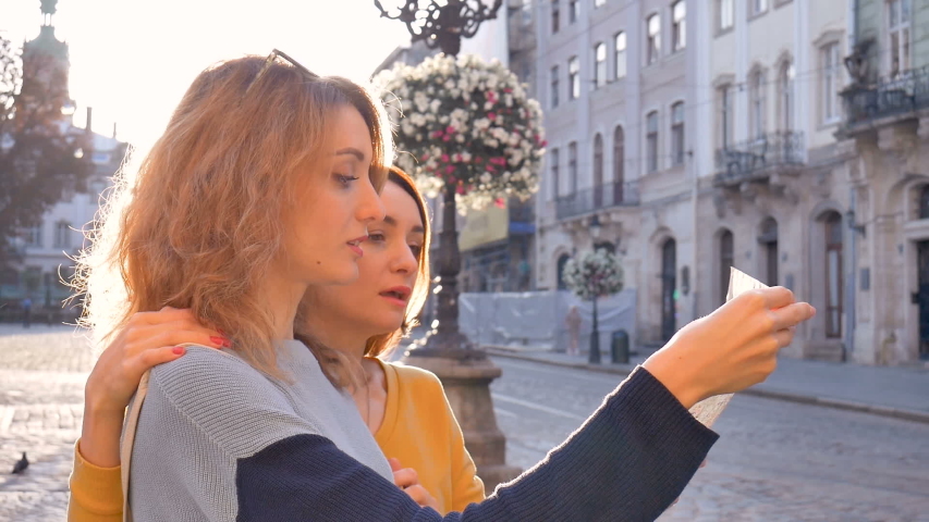 Happy young tourists holding a paper map of ancient European city early in the morning on empty square and pointing on some sightseeing place